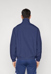 Námornícka vetrovka s vysokým golierom, elastickým lemom a bielymi bočnými pásmi. Vyrobená z ľahkej látky, má hladkú textúru.