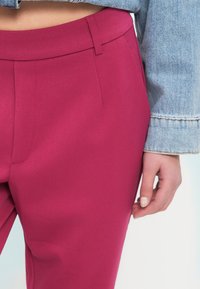 Εφαρμοστά, ψηλόμεσα παντελόνια από έντονο φούξια ύφασμα, με λεία υφή, πιέτες μπροστά και διακριτικές πλαϊνές τσέπες.