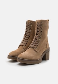 Botas de tobillo de ante marrón con punta cuadrada, parte delantera con cordones, ojales dorados y tacón grueso. Presenta una suela texturizada y detalles de costura.