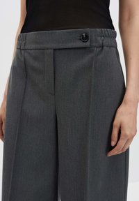 Primo piano di una persona che indossa pantaloni larghi grigio scuro con una vita bottone e una maglietta nera, in piedi davanti a uno sfondo semplice.