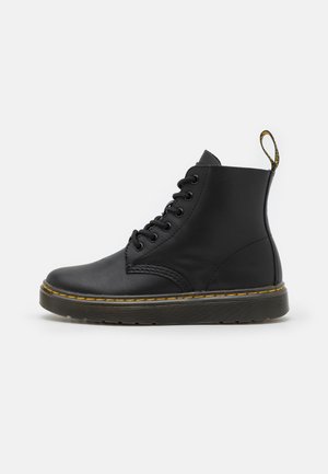 Bottine en cuir noir avec huit œillets, couture jaune sur la semelle et languette au talon sur fond blanc.