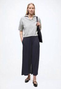 Hall hallatud poolkaelusega särk, laiad tumesinised püksid ja mustad madalad kingad. Modell hoiab musta käekotti, poseerib valge ühevärvilise tausta ees.