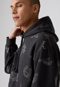 Zwarte hoodie met witte geborduurde ontwerpen, waaronder bloemen, hoefijzers en dieren. Bevat een trekkoordkap en heeft een zachte stoftextuur.