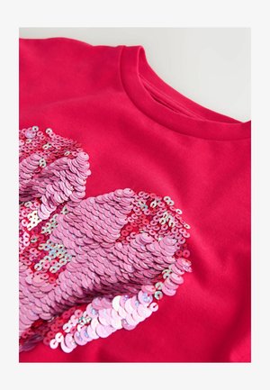 Punainen puuvillainen collegepaita, jossa on sydänkuvio, koostuen päällekkäisistä vaaleanpunaisista paljeteista, jotka luovat teksturoidun, hohtavan vaikutelman.