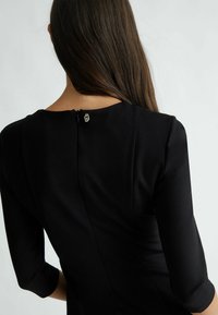 Abito nero con scollatura tonda, maniche a tre quarti e una cerniera sul retro con un dettaglio decorativo di chiusura. Tessuto dalla consistenza liscia.