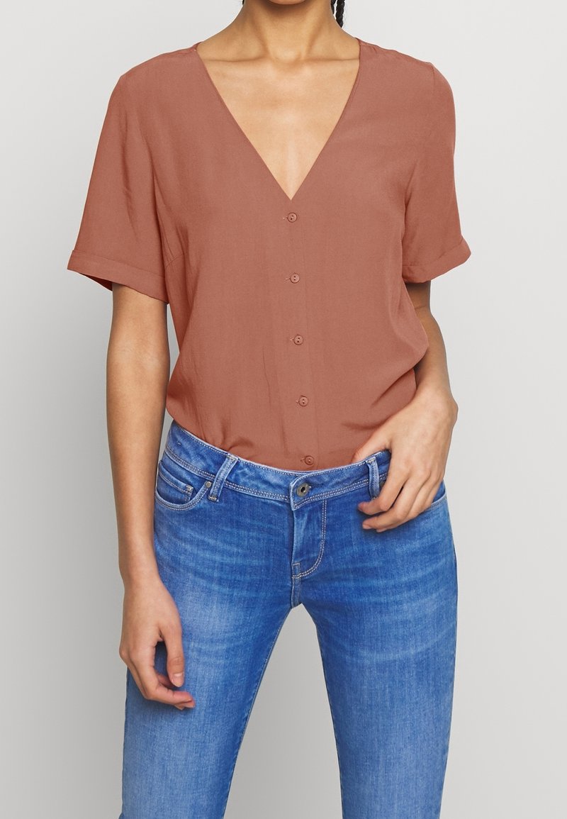 Femme portant un chemisier marron à manches courtes boutonné et un jean bleu en denim, une main posée près de la poche.