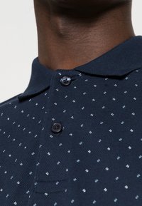 Detailní záběr na tmavou pokožku krku, který je oblečený v námořnicky modré košili s malými bílými a světle modrými tečkami a dvěma tmavými knoflíky zapnutými.