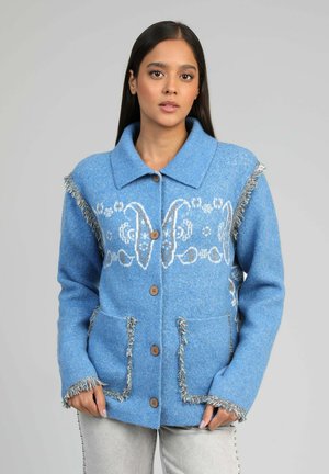 Femme portant une veste en laine bleue boutonnée avec des motifs blancs, une bordure à franges et des poches devant, se tenant devant un fond gris.