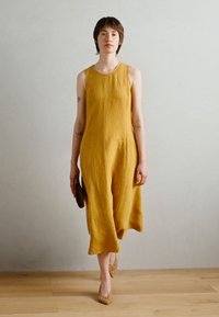 Sennepsgul lin sleeveless kjole med løs passform og kne-lengde nederkant; paret med en brun clutch og åpne, vevde sko.