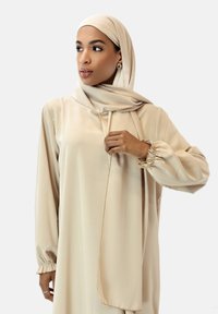 Abito beige a maniche lunghe e hijab abbinato, realizzato in tessuto liscio. Maniche a sbuffo con polsini elastici, che evidenziano un delicato accento dorato.