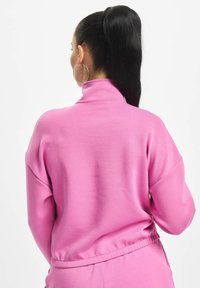 Felpa rosa con colletto alto e vestibilità rilassata. Texture liscia, spalle scese e vita elastica. Orecchini a cerchio argentati visibili.