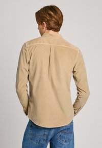 Beige corduroy shirt met lange mouwen, met een rugplooi detail en knoopsluiting bij de manchetten, gecombineerd met een blauwe spijkerbroek met achterzakken.