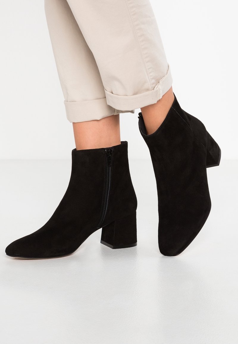 Bottes noires en daim jusqu'à la cheville avec talons carrés et fermetures éclair latérales portées avec un pantalon beige retroussé.