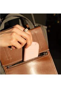 Estuche rosa etiquetado como "Rosa Brillante" parcialmente insertado en un bolso marrón de textura suave y cremallera plateada. Mano extendiéndose hacia el estuche.