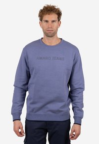 Hombre con cabello rizado que lleva una sudadera azul con el texto "AMARO JEANS", de pie contra un fondo claro y liso.