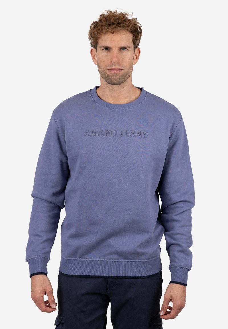 Hombre con cabello rizado que lleva una sudadera azul con el texto "AMARO JEANS", de pie contra un fondo claro y liso.