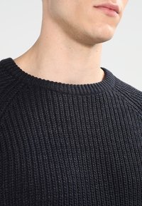 Mörkblå ribbad stickad tröja med rund hals och textilstruktur. Visar en närbild av kragen och axelområdet.