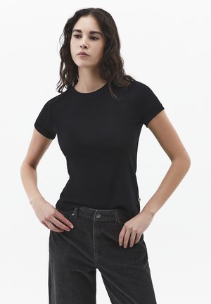 Femme portant un t-shirt noir moulant à manches courtes et un pantalon en velours côtelé noir, debout les mains sur les hanches devant un fond blanc.