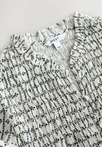 Bluza albă cu model în dungi florale negre, textură de material adunat, decolteu cu volane și etichetă a mărcii "NEXT" în interiorul gulerului.