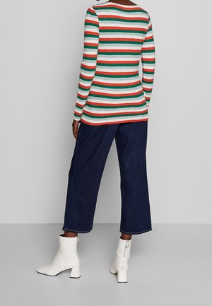 Personne portant une chemise à manches longues rayée horizontalement rouge, verte et blanche, un pantalon large foncé et des bottines blanches avec fermetures éclair.