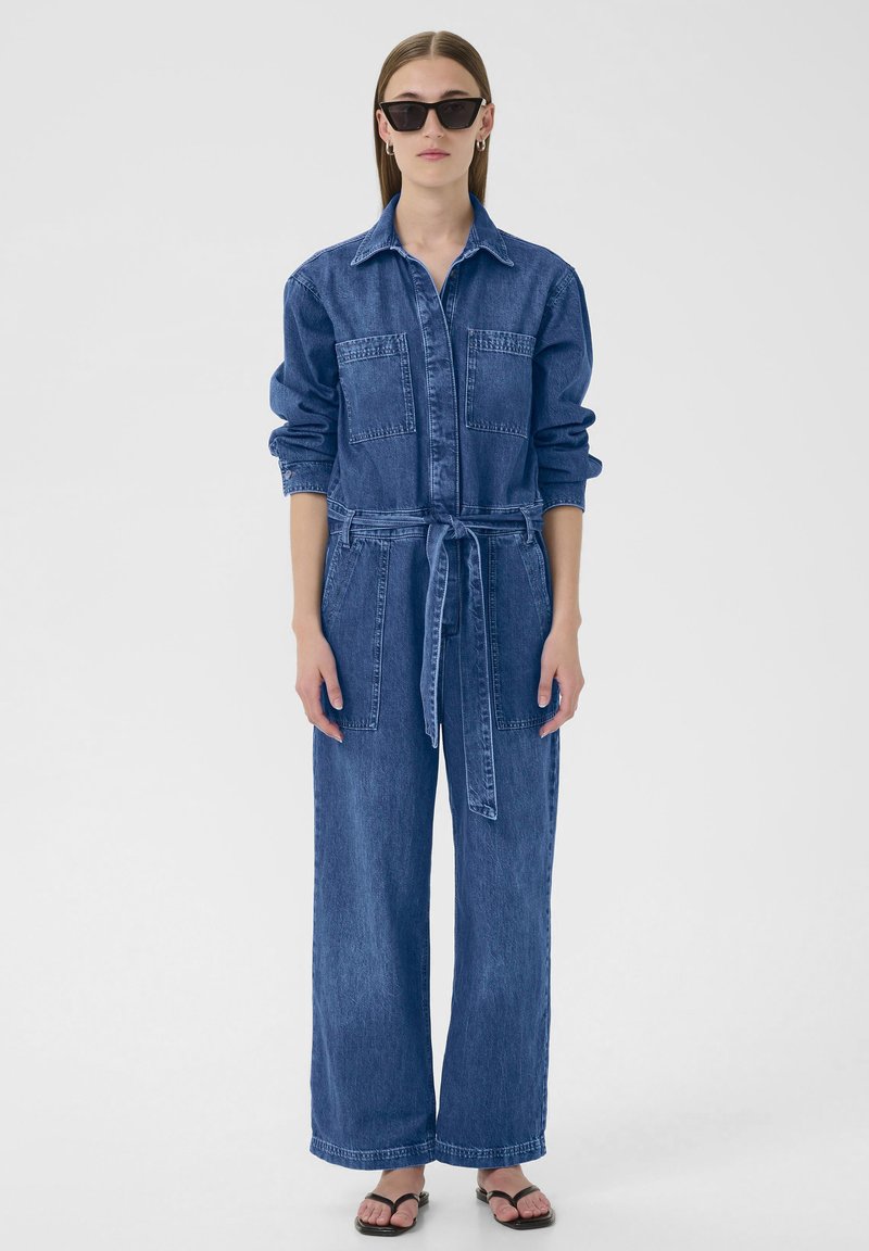 Jeansjacka med lång ärm, två bröstfickor, midjeband och vida ben i en mellanblå tvätt; bärs med svarta solglasögon och sandaler.