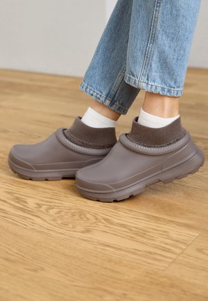 Brune gummisko uden snørebånd med en strikket overdel, struktureret krave og kraftig sål, vist på et trægulv ved siden af lyse denim.