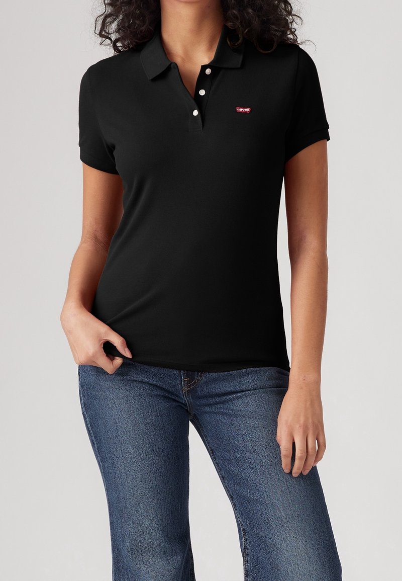 Femme portant un polo noir cintré avec des boutons blancs et un petit logo rouge Levi's, associé à un jean denim bleu.