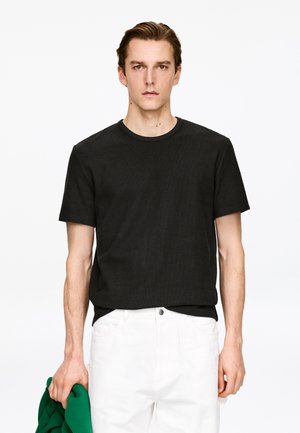 Sort ribstrikket t-shirt med korte ærmer og rund hals, kombineret med hvide bukser. Modellen holder et grønt stykke tøj i hånden.