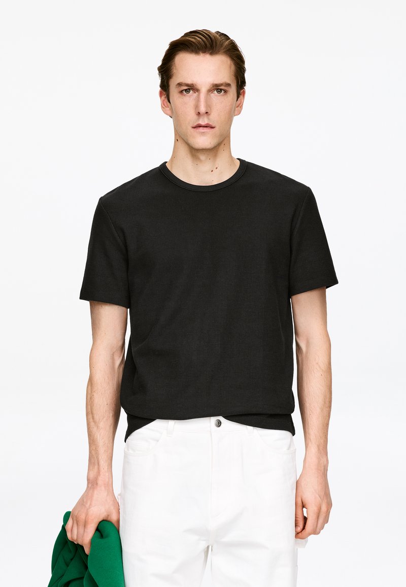 T-shirt noir à manches courtes côtelées avec un col rond, associé à un pantalon blanc. Le mannequin tient un vêtement vert à la main.