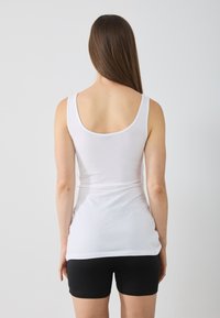 Bela bombačasta majica na naramenice z zaobljenim izrezom na hrbtu, nosila z črnimi kratkimi hlačami. Majica ima oprijet kroj in gladko teksturo.