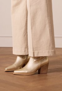 Gold leather ankle boots with a pointed toe and block heel, featuring textured accents and a smooth finish, paired with cream wide-leg pants.