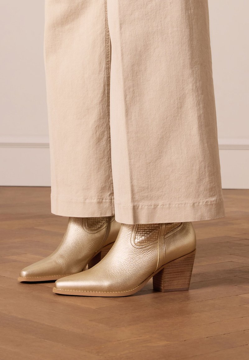 Gold leather ankle boots with a pointed toe and block heel, featuring textured accents and a smooth finish, paired with cream wide-leg pants.