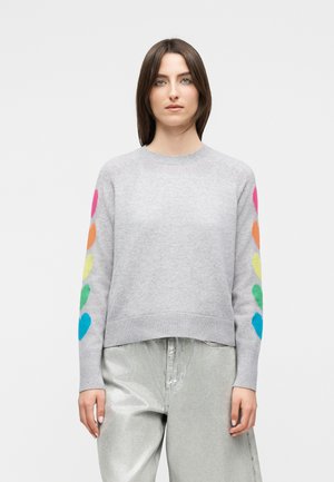 Žena v svetlosivom svetri s viacfarebnými srdcovými tvarmi na oboch rukávoch a voľných lesklých strieborných nohaviciach, stojaca nehybne.