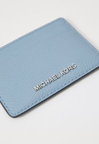 Jasnoniebieski skórzany portfel na karty z teksturowaną powierzchnią i srebrnym logo "MICHAEL KORS". Posiada wiele miejsc na karty i przeszywane krawędzie.