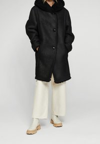 Abrigo negro con capucha de peluche, que presenta botones grandes, bolsillos laterales y un acabado texturizado. Combinado con pantalones de pierna ancha color crema y botas claras.