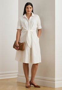 Balta lina krekla kleita ar mezglu priekšpusē, īsām piedurknēm un sānu kabatām. Kombinēta ar brūnu somiņu un smailu purngalu augstpapēžu kurpēm.