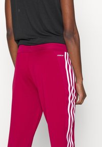 Bordeauxfarbene Sporthose mit weißen Seitenstreifen und "AEROREADY"-Logo am Bund. Weicher, elastischer Stoff für Komfort und Bewegungsfreiheit.