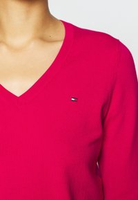 Maglione rosa a scollo a V realizzato in tessuto a maglia. Presenta un piccolo logo sul petto. La texture appare liscia e morbida.