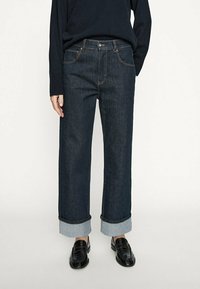 Mörkblå denimjeans med vida ben och uppvikt fåll. Har fem fickor, kontrasterande sömmar och en framknäppning.