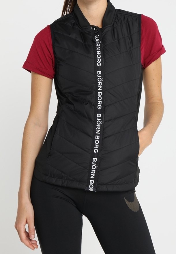 Gilet sans manches matelassé noir avec fermeture éclair à l'avant et bande blanche de marque au centre. Porté sur une chemise rouge à manches courtes.