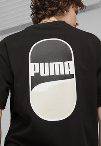 Juodas medvilnės marškinėliai su dideliu ovaliu logotipu ant nugaros, pilkos ir kreminės spalvų, su užrašu "PUMA" ryškiais baltais rašmenimis.