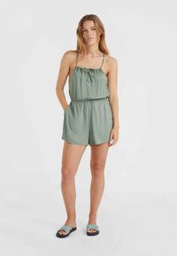 Un mono verde sin mangas con cintura fruncida y detalle de lazo en la parte delantera. Se lleva con sandalias de tiras trenzadas, que presentan una parte superior texturizada y suela de goma.