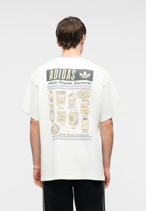 Man draagt een wit Adidas T-shirt met een retro supermarktgrafiek met levensmiddelen en tekst, gezien van achteren tegen een effen achtergrond.