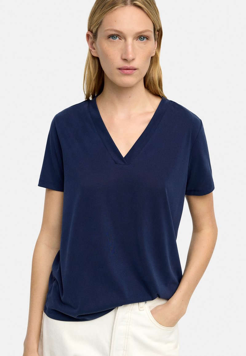 Granatowa koszulka z dekoltem w serek, uszyta z miękkiego materiału, z krótkimi rękawami i luźnym krojem, zestawiona z jasnymi spodniami.