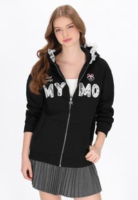 Sudadera negra con cremallera y capucha con borde de encaje, con la inscripción "MY MO" en letras blancas y un pequeño emblema rosa. Combinada con una falda gris plisada.