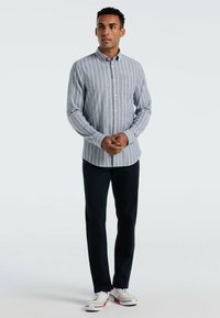 Lichtgrijs overhemd met witte verticale strepen, voorzien van lange mouwen en een enkele borstzak, gecombineerd met donkere broek en sneakers.