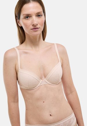 Soutien-gorge beige orné de dentelle avec une texture douce, des bonnets légèrement rembourrés et des bretelles ajustables, présentant des motifs floraux et des bords festonnés.
