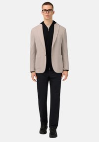 Ljusgrå kavaj med två framfickor över en svart zip-up hoodie, kombinerad med mörka byxor och svarta sneakers. Tydlig och strukturerad passform.