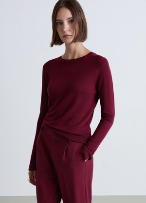 Woman with short brown hair wearing a long-sleeve burgundy top and matching pants, standing with one hand in her pocket.