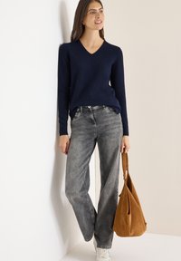 Marineblauer V-Ausschnitt-Pullover, graue weit geschnittene Jeans und weiße Sneakers. Hält eine Handtasche aus braunem Wildleder. Einfaches Design mit strukturierter Oberfläche.